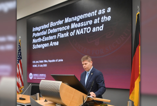 Raitis Tiliks stands at a podium delivering his alumni scholar presentation at the Marshall Center, with a large screen behind him displaying the title of his research on integrated border management and deterrence on NATO’s Northeastern flank