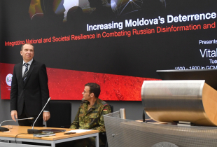 Vitalie Armașu stands at the front of a conference room next to two seated individuals, with a presentation slide behind him titled 'Increasing Moldova's Deterrence Potential.' An American and German flag are visible on either side of the screen.