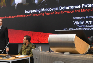 Vitalie Armașu stands at the front of a conference room next to two seated individuals, with a presentation slide behind him titled 'Increasing Moldova's Deterrence Potential.' An American and German flag are visible on either side of the screen.