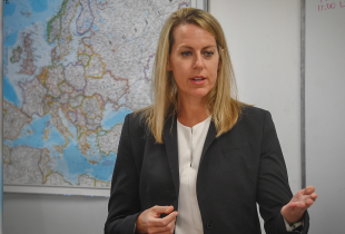 A PLTCE faculty member speaks to participants during a classroom session, gesturing with one hand while standing in front of a map of Europe and a whiteboard.