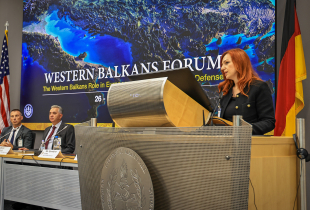 Gjeraqina Leka, assistant professor at the University of Tetova, speaks at the podium during the Western Balkans Forum at the Marshall Center in Garmisch-Partenkirchen, Germany. Behind her, a screen displays the event title alongside the U.S. and German flags. Three panelists are seated at the table to her left.