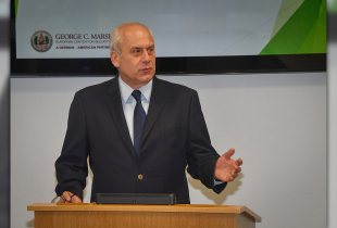 A man stands behind a podium, raising one hand in emphasis as part of a presentation, while in front of a monitor displaying the Marshall Center logo.