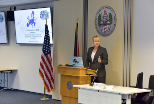 Blonde woman speaks behind a podium with US and German flag behind her. 