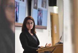 A woman with glasses stands at a podium giving a presentation, with a projection screen visible in the background.