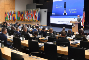 A large group of participants sit in an auditorium facing a stage where a speaker addresses them at a podium. Behind the speaker, a large screen displays the Program on Applied Security Studies dates and participant information, with rows of international flags lining the wall.