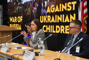 Three speakers sit at a panel table during the Program on Applied Security Studies. A large screen behind them reads “War of Russia Against Ukrainian Children,” and an American flag stands to the right.