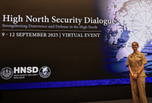 A person in a tan U.S. Navy uniform stands smiling in front of a large screen that reads “High North Security Dialogue” beside a map of the Arctic.