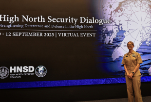 A person in a tan U.S. Navy uniform stands smiling in front of a large screen that reads “High North Security Dialogue” beside a map of the Arctic.