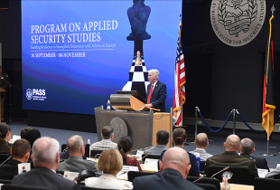 PASS participants sit in a large auditorium as Director Barre Seguin speaks at a podium beside a large course banner and rows of national flags.