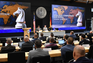 Speaker at a podium addresses participants during the Marshall Center’s “Strategic Competition and Russia” course. A panel sits beside him, with large screens showing a world map and chess knight graphic, and international flags displayed along the wall. 