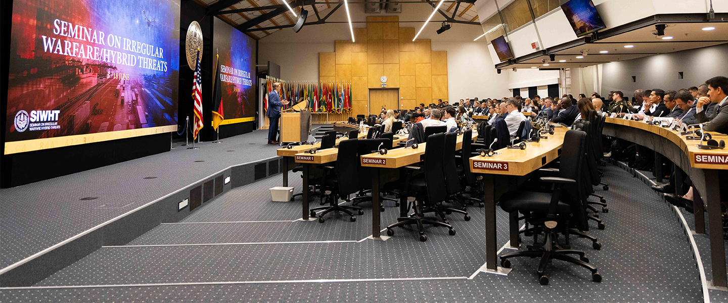 A wide-angle view of the plenary room at the George C. Marshall Center during the Seminar on Irregular Warfare/Hybrid Threats. A speaker addresses a full auditorium, with participants seated at curved desks labeled “Seminar 1,” “Seminar 2,” and “Seminar 3.” Two large screens display the seminar title and dates, framed by U.S. and German flags and a backdrop of international flags.