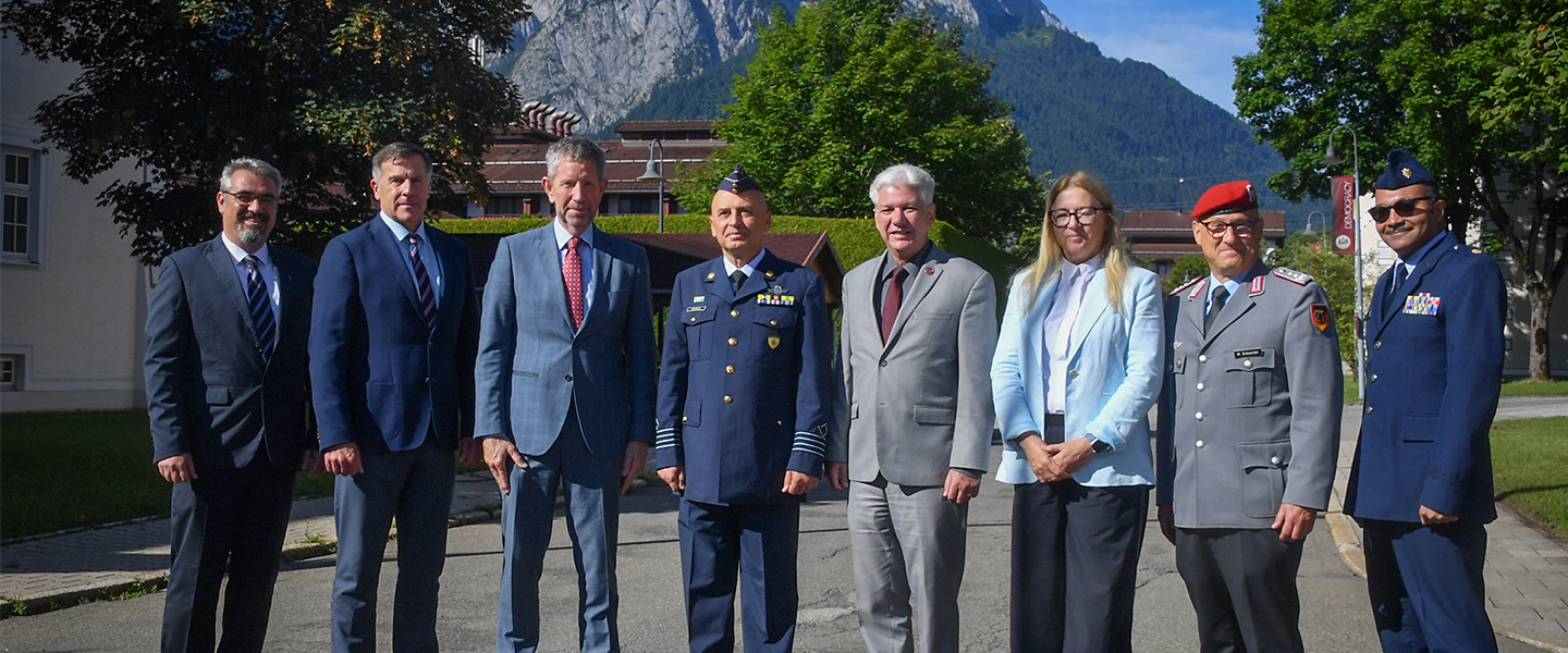 Eight people stand in a line outdoors, dressed in formal business and military attire, posing for a group photo. Behind them are trees, buildings, and a mountain range under a clear blue sky.