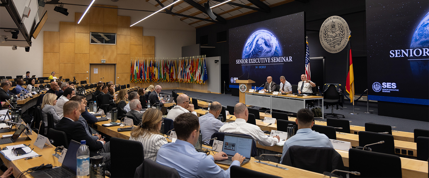 Participants attend a plenary session of the Senior Executive Seminar at the George C. Marshall Center. A panel of speakers sits on stage beneath a large screen displaying the seminar title, “Senior Executive Seminar, 14–18 July.” The auditorium is filled with attendees seated at desks, many using laptops, while a wall of international flags lines the back of the room. U.S. and German flags are displayed near the speakers.