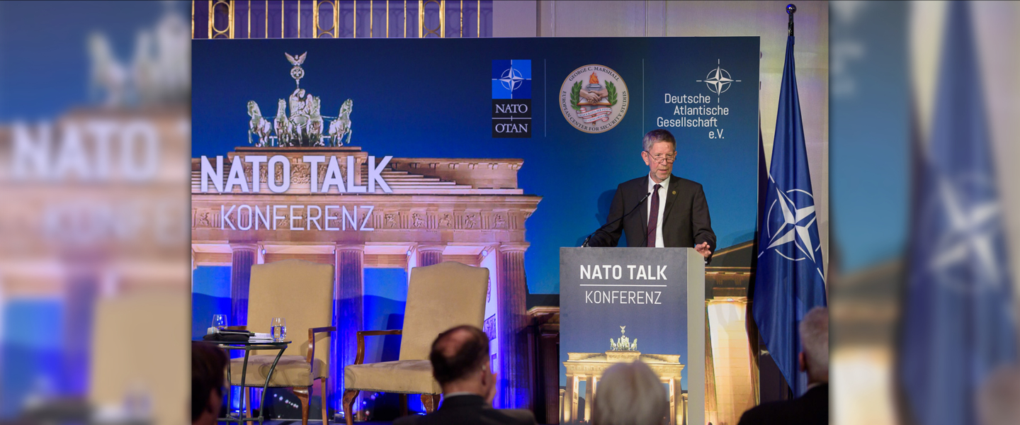 A man presents behind a podium in front of a NATO Talk 2025 backdrop.