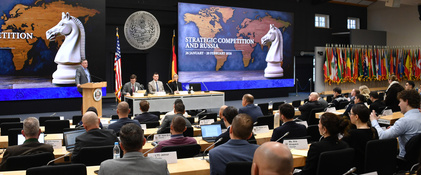 Speaker at a podium addresses participants during the Marshall Center’s “Strategic Competition and Russia” course. A panel sits beside him, with large screens showing a world map and chess knight graphic, and international flags displayed along the wall. 