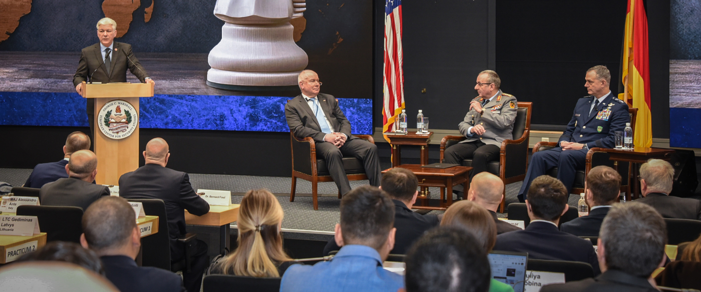 Senior leaders speak during a panel at the Marshall Center beneath U.S. and German flags, with a large chess knight and world map graphic displayed behind them during the Strategic Competition and Russia course, January 2026.